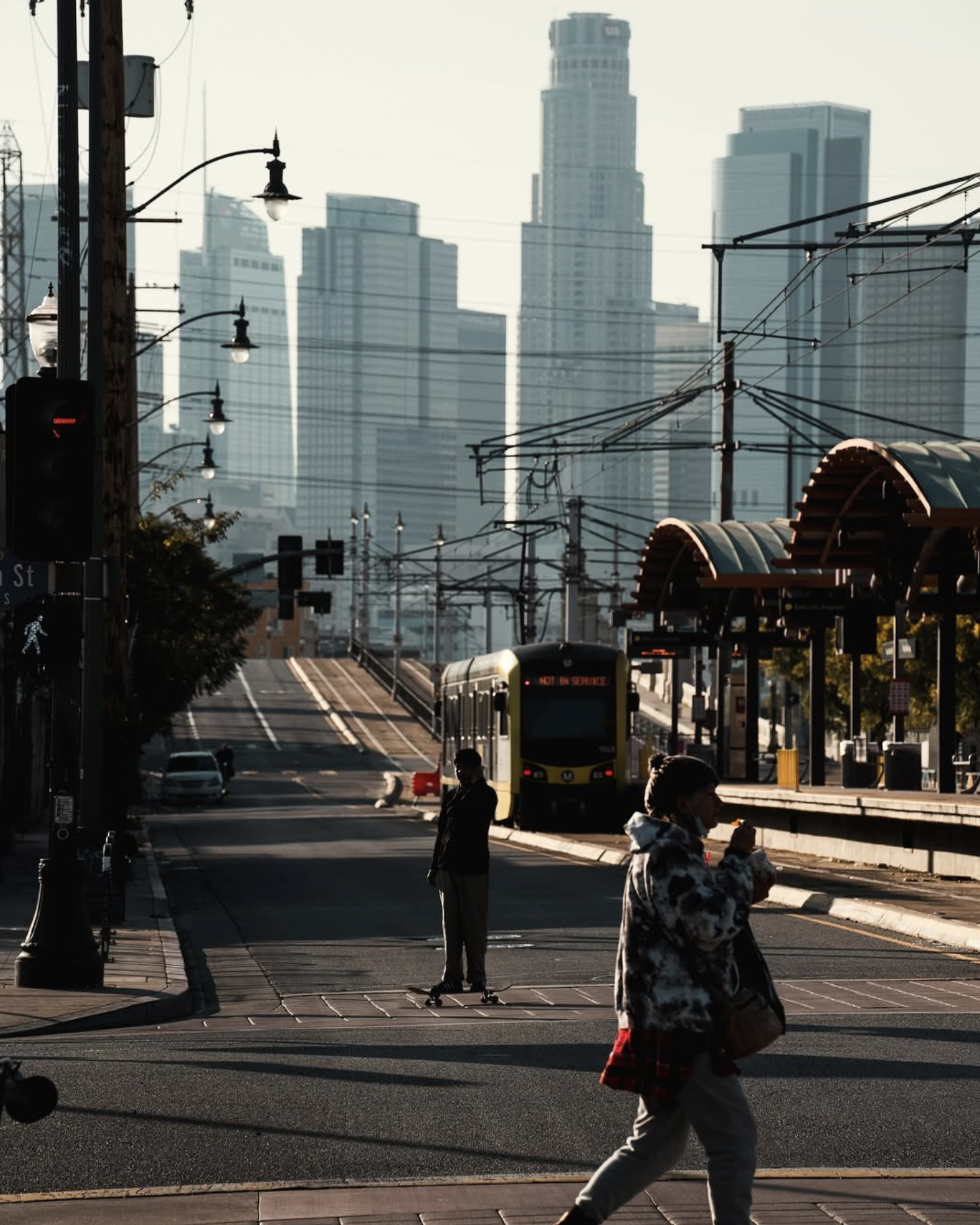 A skateboarder crosses the street in front of a Metro light rail train, downtown Los Angeles skyline behind
